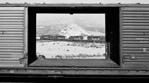 A black and white image of an open train car that reveals another train through it.